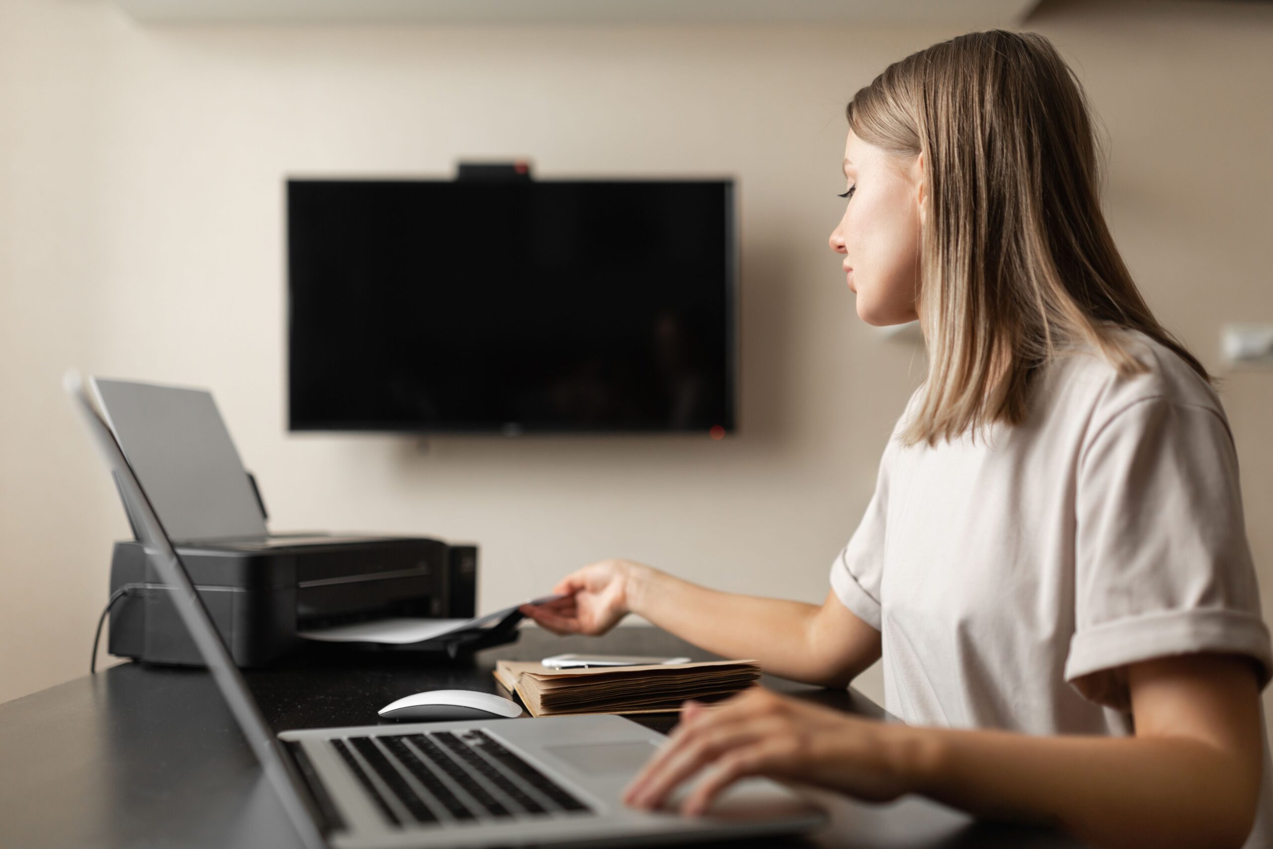 A woman sitting at a desk operates a laptop while feeding paper into a printer. A television is mounted on the wall behind her.