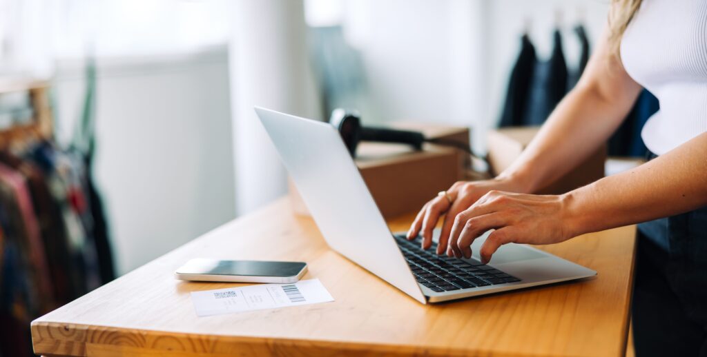 A person types on a laptop at a wooden desk, with a smartphone and shipping label nearby, suggesting an online business or retail setup.