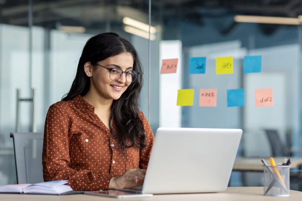 Smiling woman in a polka dot shirt works on a laptop, surrounded by colorful sticky notes, in a modern office setting.