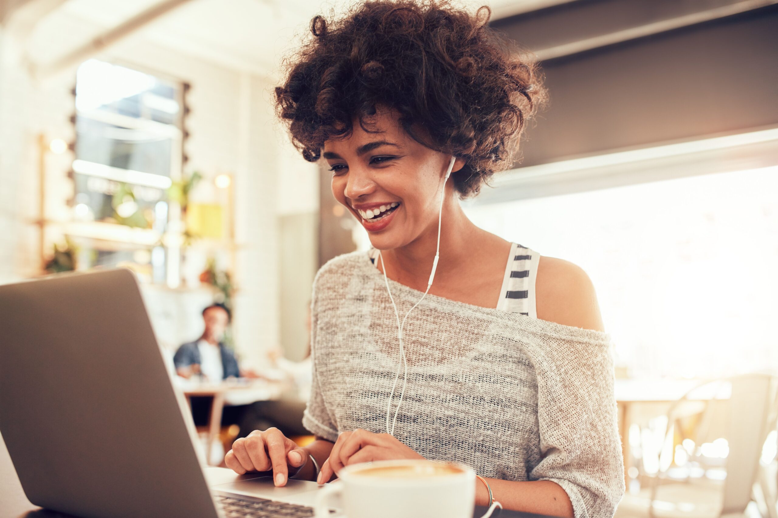 Young woman in a cozy café smiles while using a laptop with headphones. A coffee cup is visible, highlighting a relaxed work or study atmosphere.