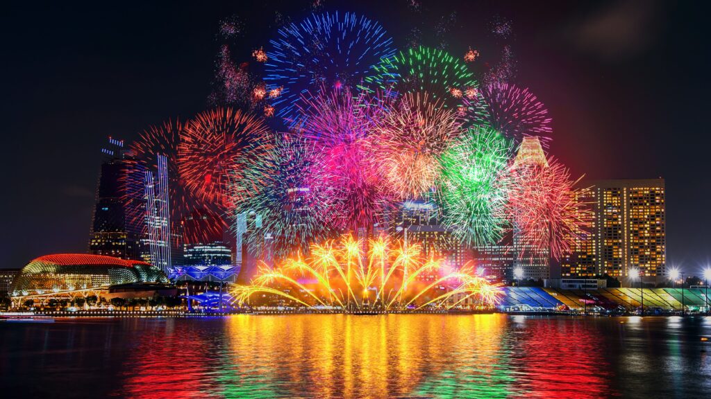 Vibrant fireworks illuminate the night sky above a city skyline, reflecting in the water below, celebrating a festive occasion.