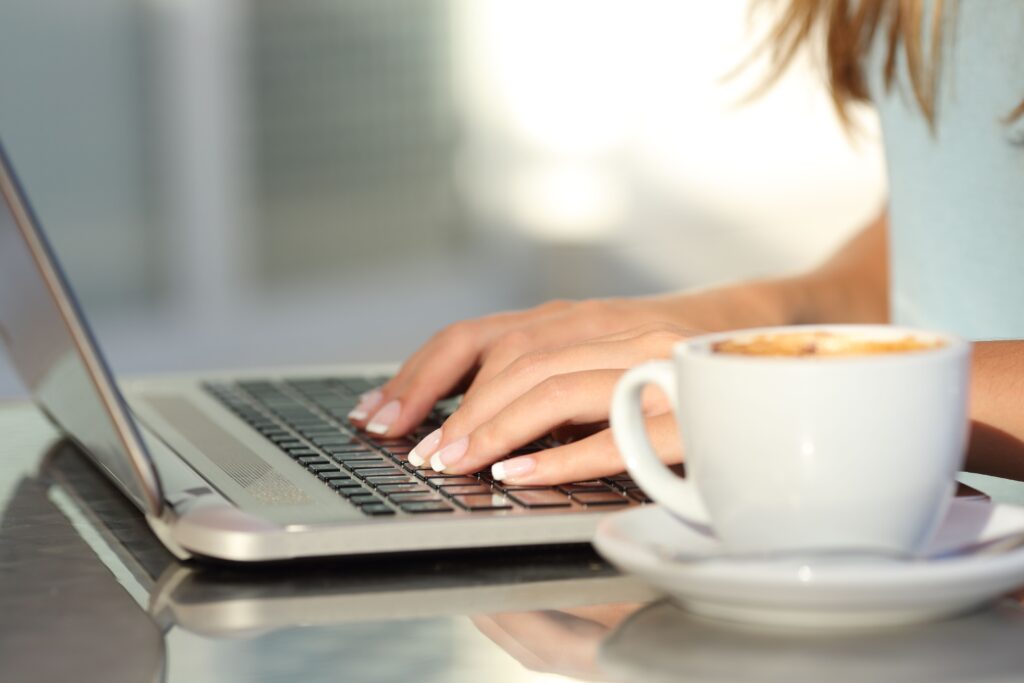 A person types on a laptop in a bright setting, with a coffee cup nearby, suggesting a casual work or study environment.