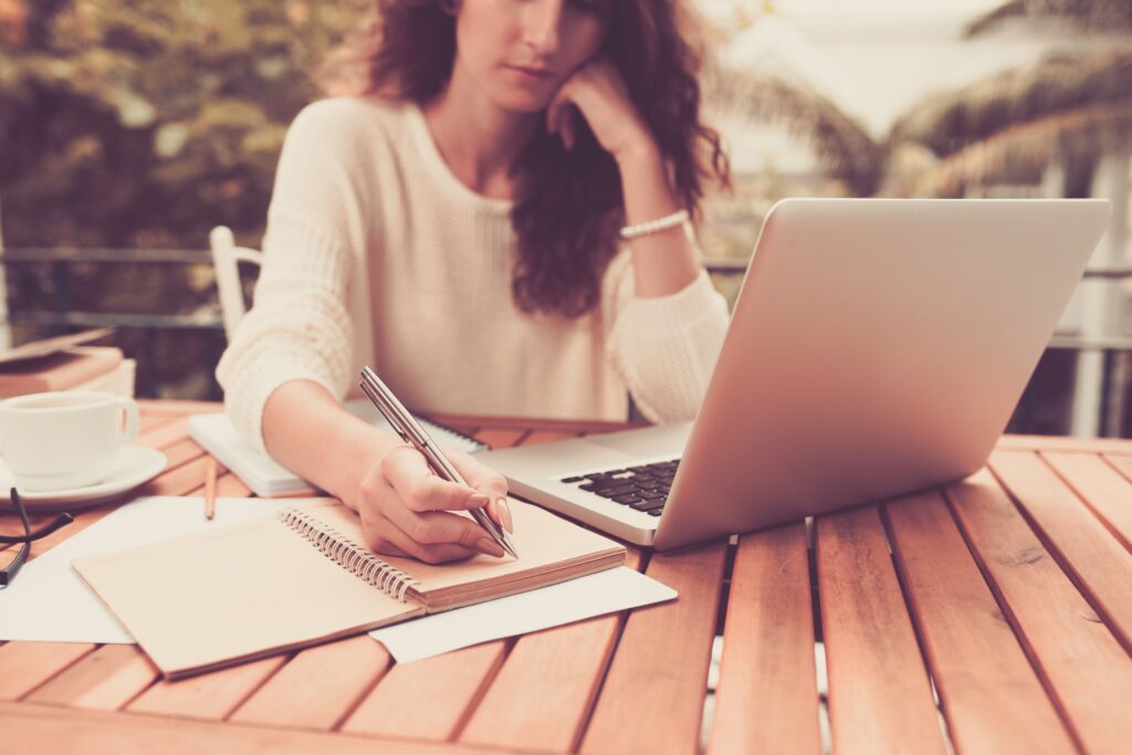 Woman writing in a notebook while working on a laptop at an outdoor table, surrounded by papers and a coffee cup. Ideal for illustrating a productive workspace.