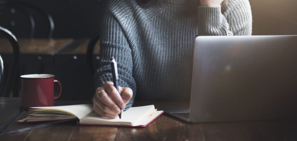 A person in a gray sweater writes in a notebook while seated at a wooden table, with a laptop and a red mug nearby. This reflects a focused, creative workspace.
