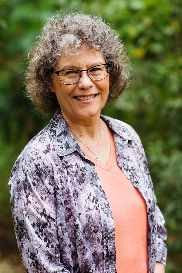 Smiling woman with curly hair, wearing glasses and a patterned shirt, stands outdoors against a blurred green background.